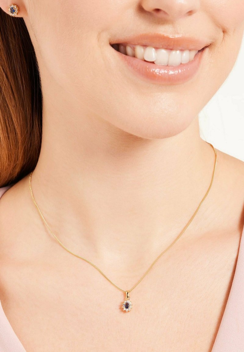 Collier en or avec un pendentif rond comportant une pierre précieuse bleue centrale entourée de petites pierres claires, porté sur la peau nue.