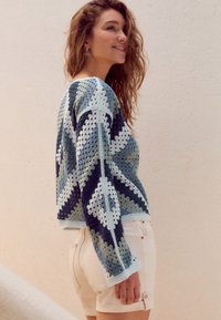 Sweter na szydełku w odcieniach niebieskiego, zielonego i białego z geometrycznymi wzorami. Połączony z białymi szortami. Długie rękawy, zaokrąglony dekolt.