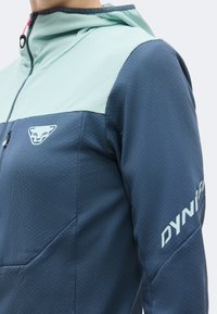 Κοντινό πλάνο ενός ατόμου που φοράει δίχρωμο μπλε και βεραμάν μπουφάν Dynafit με φερμουάρ στον γιακά και λογότυπο στο στήθος και στο μανίκι.