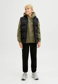 Gilet trapuntato nero sopra una felpa verde oliva con logo, abbinato a pantaloni sportivi neri e scarpe da ginnastica grigio chiaro. Sfondo semplice.