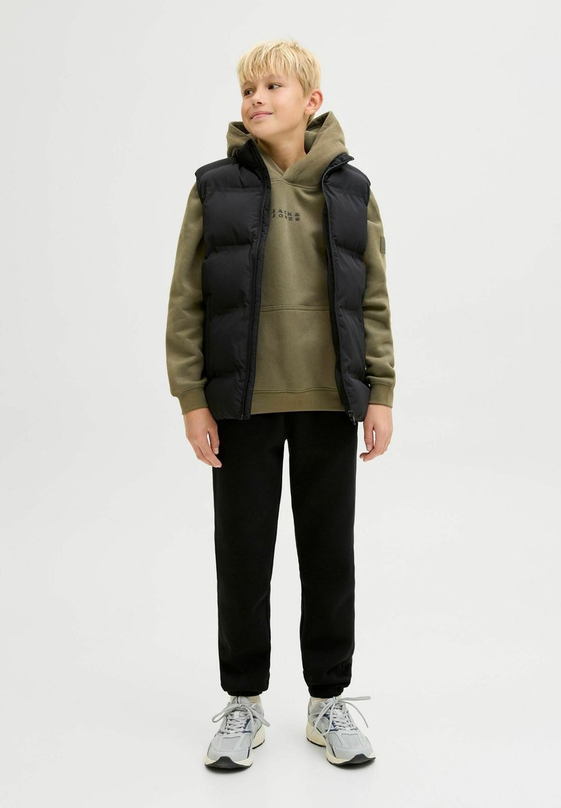 Gilet trapuntato nero sopra una felpa verde oliva con logo, abbinato a pantaloni sportivi neri e scarpe da ginnastica grigio chiaro. Sfondo semplice.