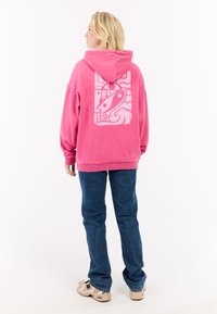 Haut à capuche surdimensionné fuchsia avec un grand motif blanc au dos. Associé à un jean bleu et des baskets beige, avec une texture douce.