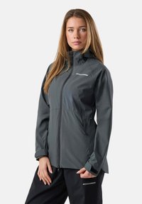 Mladá žena so svetlohnedými rovnými vlasmi, oblečená v tmavosivej kapucňovej bunde na zips a čiernych nohaviciach s reflexnými logami, stojaca na bielom pozadí.