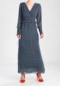 Długa, granatowa sukienka w kwiaty z lekkiego materiału, z głębokim dekoltem w kształcie V i marszczoną talią. Stylizowana z czerwonymi, wysokimi szpilkami, z długimi rękawami.