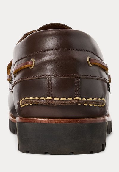 Brun skinnbåt-sko med mokasinsting, skinnsnøringer trukket gjennom metalløyer og en svart gummisåle med knotter, sett bakfra.
