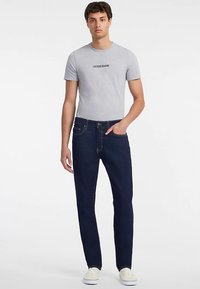 Grå bomulds-T-shirt med "GUESS JEANS" logo, kombineret med mørkeblå denimjeans med gule syninger, slim fit og lommer. Hvide sneakers.