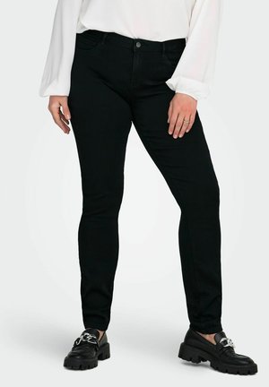 Femme portant un pantalon noir slim, un chemisier blanc à manches longues et des mocassins noirs épais avec des chaînes argentées, debout, mains détendues.