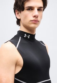 Νέος άνδρας που φοράει αμάνικη μαύρη συμπιεστική μπλούζα Under Armour με ψηλό γιακά και λευκή ραφή, ουδέτερο φόντο.