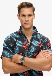 Camisa de manga corta negra con patrones de hojas tropicales en vibrantes tonos azules y rojos; el modelo lleva un reloj con esfera negra y correa de piel.