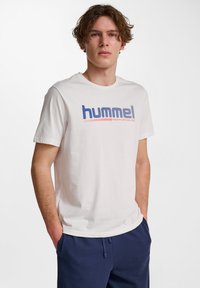 Vit bomullströja med en blå och röd "hummel PERFORMANCE" grafik. Klassisk rundhalsdesign, korta ärmar, avslappnad passform.