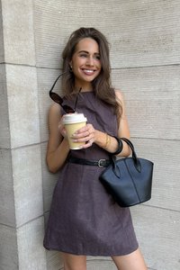 Fittovaná, bezrukávová hnedá ľanová šaty s opaskom, skombinované s malou čiernou štruktúrovanou taškou. Drží pohár s kávou so zakrývačom.