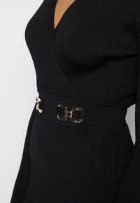 Schwarzes, geripptes Kleid mit tiefem V-Ausschnitt und langen Ärmeln, das mit einem goldfarbenen Schnallen-Gürtel in der Taille für zusätzlichen Akzent versehen ist.