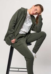 Junge Frau in olivgrünem Trainingsanzug und weißen Sneakers sitzt auf einem Hocker, lehnt den Kopf auf die Hand und lächelt leicht vor einfarbigem Hintergrund.