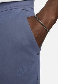 Nike blauwe trainingsbroek heeft een zijzak, mat logo en een gladde stof met een subtiele textuur. Zilveren kettingarmband gedragen aan de hand.