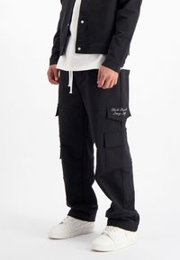 Zwarte cargobroek met zakken, textuurstof, taille met trekkoord en geborduurd logo detail. Gemaakt in combinatie met witte sneakers.