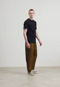 Černé polo tričko s krátkým rukávem a bílými lemy, olivově zelené kalhoty a šedé tenisky s bílou podešví. Model stojí před neutrálním pozadím.