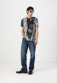 Kortslemet svart-hvitt mønstret t-skjorte, med abstrakte design; kombinert med blå denimjeans og svarte sko. Modellen har på seg grå solbriller.
