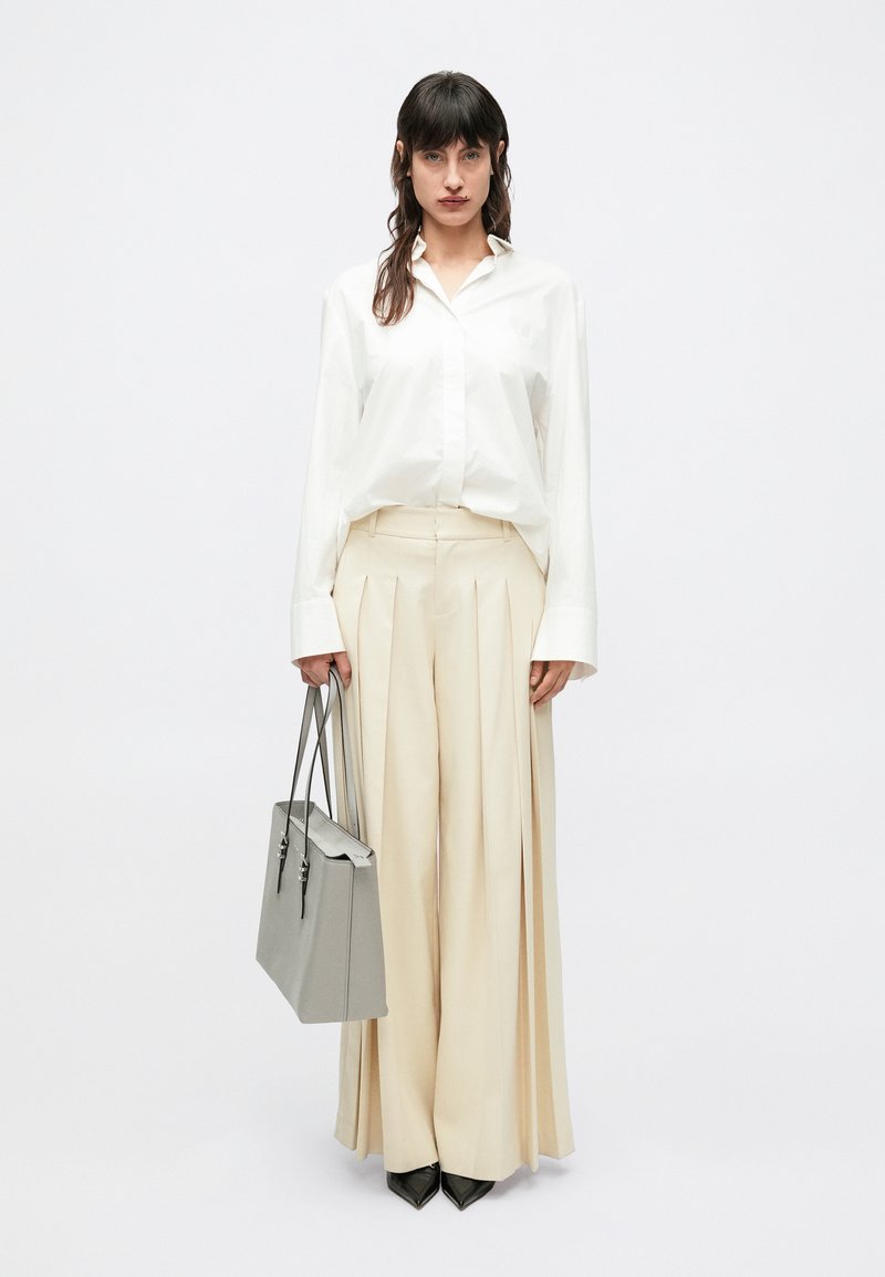 Λευκό πουκάμισο με κουμπιά, μπεζ πλισέ παντελόνι, μαύρα μυτερά τακούνια και γκρι δομημένη τσάντα χειρός. Καθαρές γραμμές και αντιθετικά χρώματα.