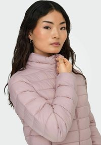 Heledad roosa puffjakker, millel on tikanditega disain, kõrge kaelus ja lukuga sulgemine. Sile tekstuur ja sobiv siluett koos peente aktsentidega.