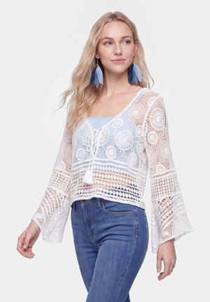 Blonde Frau mit großen blauen Federohrringen, einer weißen gehäkelten Bluse über einem hellblauen Top und blauen Jeans, die zur Seite schaut.