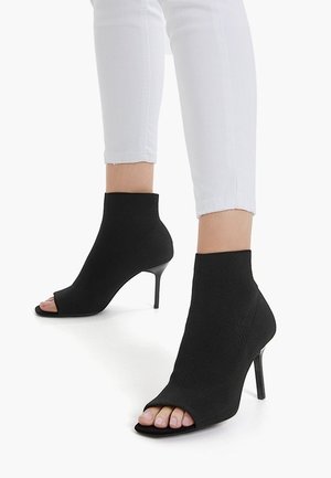 Bottines noires à bout ouvert avec talons aiguilles portées avec un pantalon court blanc, montrant les orteils nus et une texture de tissu lisse.
