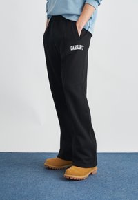 Μαύρο φόρμα παντελόνι με πλαϊνές τσέπες και λευκό λογότυπο "Carhartt". Συνδυάζεται με καφέ μπότες υψηλού προφίλ πάνω σε μπλε χαλί.