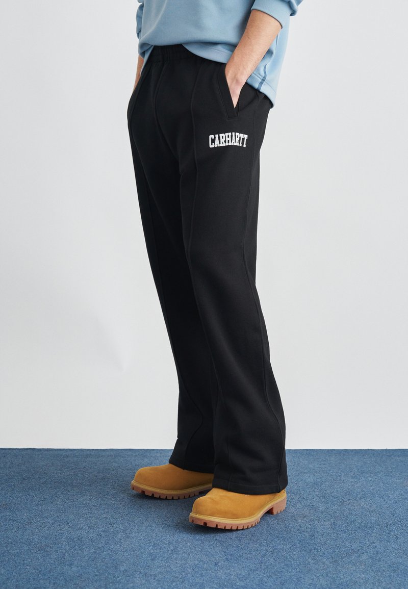 Μαύρο φόρμα παντελόνι με πλαϊνές τσέπες και λευκό λογότυπο "Carhartt". Συνδυάζεται με καφέ μπότες υψηλού προφίλ πάνω σε μπλε χαλί.