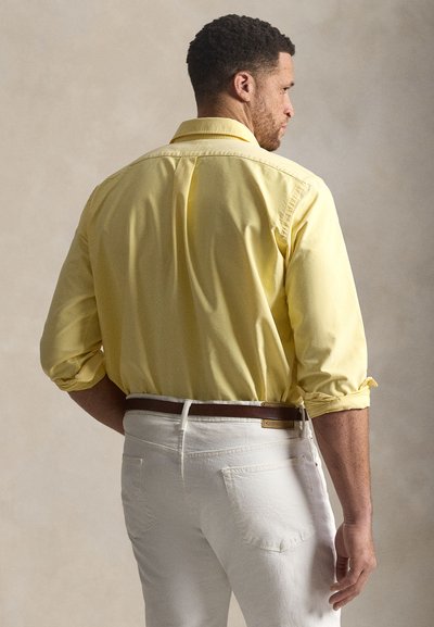 Bărbat văzut din spate purtând o cămașă galbenă cu mâneci lungi suflecate, pantaloni albi și o curea maro pe un fundal neutru.