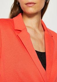 Blazer corail avec col structuré et tissu texturé, porté sur un haut noir. Présente un design épuré et ajusté avec une légère brillance.