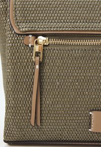 Textured olive-green material bag with brown leather accents, gold zipper, and a front pocket. Features a tassel and woven pattern.