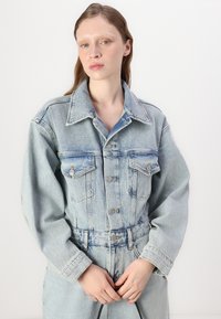Világoskék farmeroverál, galléros nyakkal, gombos elülső résszel és két mellzsebbel; laza ujjakkal és övvel ellátott derékkal rendelkezik.