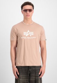 Beige kortærmet t-shirt med hvid "ALPHA INDUSTRIES" logo, sat sammen med mørke jeans og lyse solbriller, foran en ensfarvet baggrund.