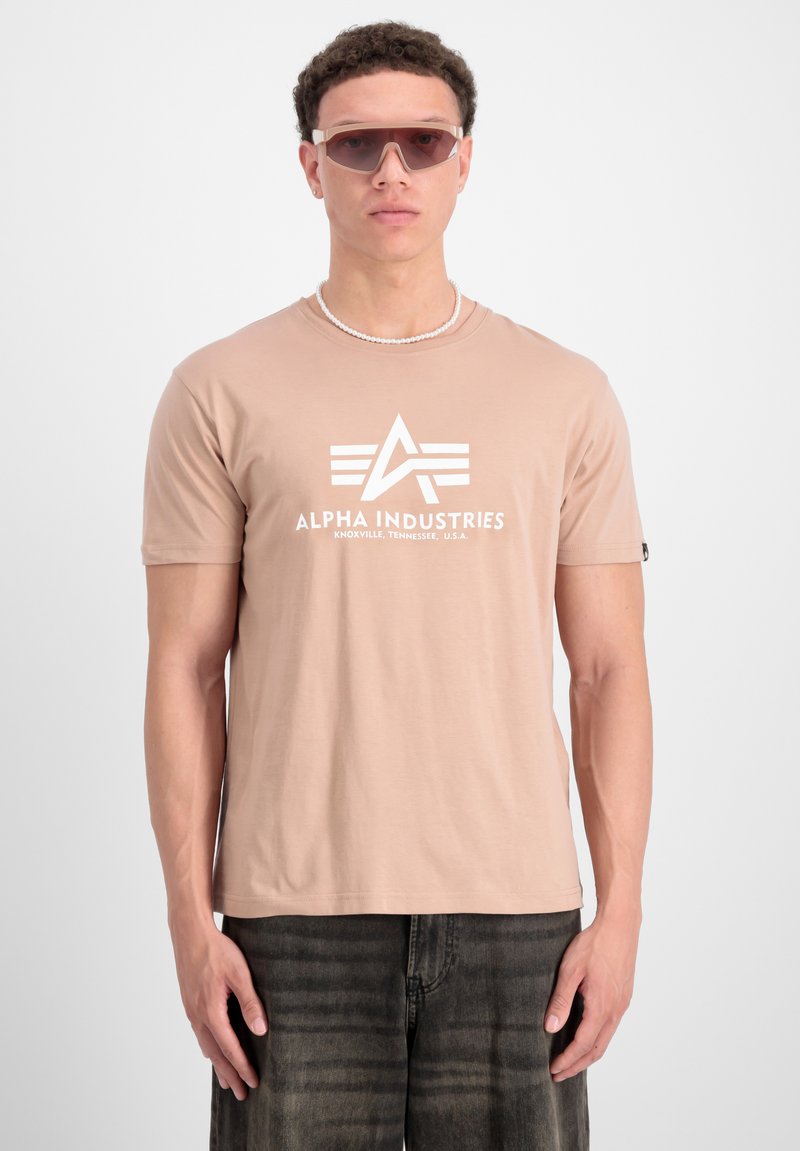 Beige kortærmet t-shirt med hvid "ALPHA INDUSTRIES" logo, sat sammen med mørke jeans og lyse solbriller, foran en ensfarvet baggrund.