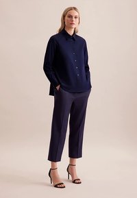 Granatowa bluzka z ostrym kołnierzykiem i guzikiem z przodu, w zestawieniu z dopasowanymi granatowymi spodniami i czarnymi sandałami na paski. Gładka faktura tkaniny.