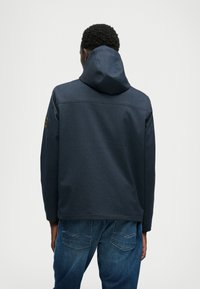 Giacca con cappuccio blu scuro realizzata in tessuto testurizzato, caratterizzata da un taglio dritto e un design semplice. Include una toppa sulla manica sinistra.