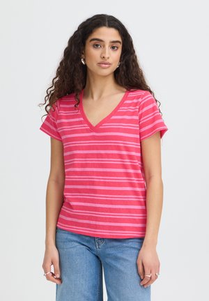 Giovane donna con capelli lunghi e ricci che indossa una maglietta a righe rosa e rosse con scollo a V e maniche corte e jeans blu, in piedi davanti a uno sfondo uniforme.