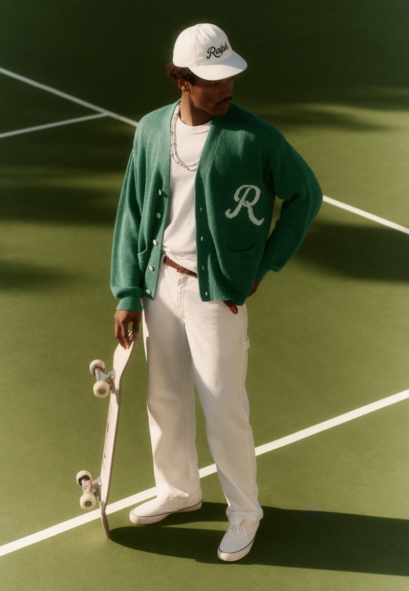 Uomo che indossa un cardigan verde e un berretto bianco che tiene in mano uno skateboard, in piedi su un campo sportivo con linee bianche.
