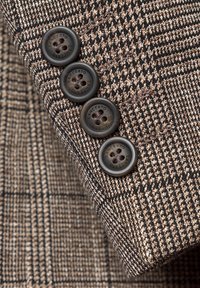 Brązowa i czarna tkanina houndstooth z czterema ciemnymi guzikami na mankiecie dopasowanej marynarki, z teksturowanym, tkanym detalem.