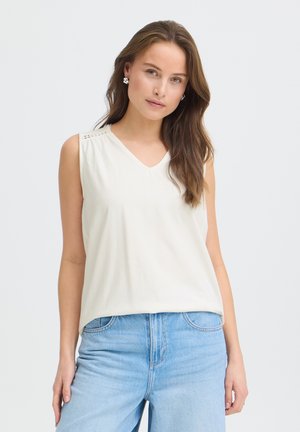 Młoda kobieta ubrana w biały, bezrękawnik z dekoltem w serek i jasnoniebieskie dżinsy, pozująca z odprężonym wyrazem twarzy na jednolitym tle.