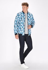 Giubbotto bomber blu con un motivo di surfisti e onde; presenta polsini a coste, chiusura con zip e tessuto leggero. Abbinato a jeans scuri.