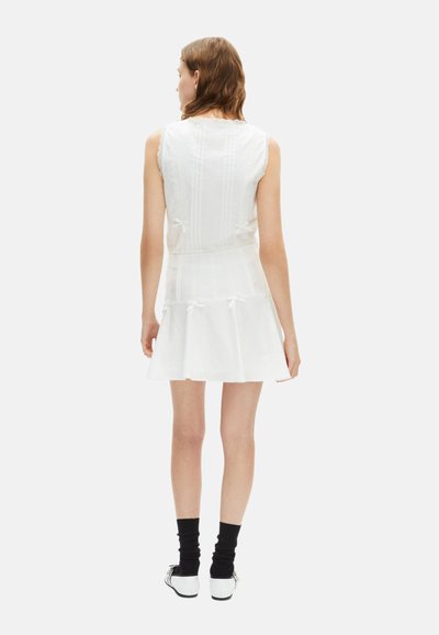 Robe blanche sans manches avec des détails en dentelle, jupe plissée et nœuds à l'arrière ; associée à des chaussettes noires et des chaussures blanches.