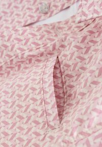 Růžová látka se vzorem geometrických tvarů, obsahující knoflík a boční kapsu. Textura je hladká a design je decentní, přesto však výjimečný.