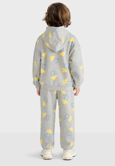 Enfant aux cheveux bruns portant un sweat à capuche gris et un pantalon avec des formes abstraites jaunes et bleues, debout de dos sur fond blanc.