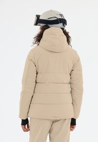 Beige pufferjacka med huva, med horisontella stygnpaneler. Svarta ärmslut, bärs med skidglasögon och hjälm.