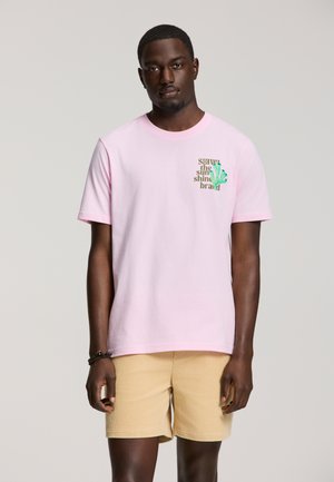 Hombre con una camiseta rosa claro con texto y gráfico, pantalones cortos beige y una pulsera negra, de pie frente a un fondo liso.