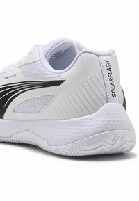 Chaussure de sport blanche avec tige en mesh et accents noirs, talon texturé et semelle à motif conçue pour la stabilité et le soutien.
