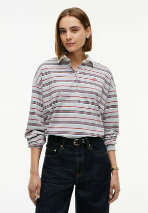 Kvinde iført en grå poloshirt med lange ærmer med røde og sorte striber og mørke jeans med sort bælte på en hvid baggrund.