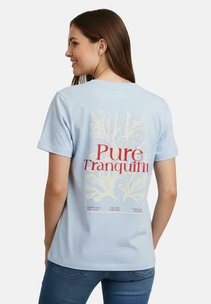 Jonge vrouw in een lichtblauw T-shirt met een koraalrode print en rode tekst "Pure Tranquility" op de achterkant, gecombineerd met een blauwe spijkerbroek.
