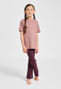 Młoda dziewczynka z warkoczami stoi boso, ubrana w fioletowawy krótkim rękawem t-shirt i ciemnofioletowe legginsy na gładkim tle.