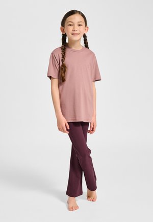 Ung pige med fletninger står barfodet iført en malva kortærmet shirt og mørkelilla leggings mod en ensfarvet baggrund.